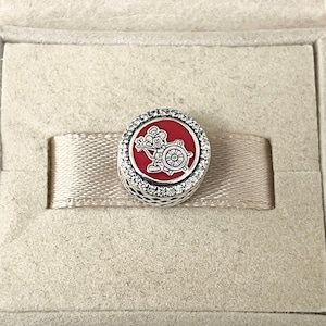 May include: A silver charm with a red enamel background featuring a cartoon mouse holding a steering wheel. The charm is surrounded by a clear crystal halo.