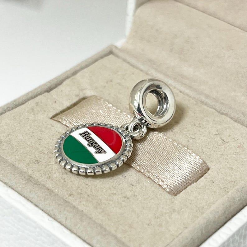 May include: Silver charm with a round enamel pendant featuring the Hungarian flag and the word "Hungary" in black lettering.
