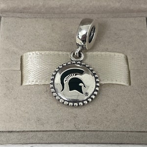 Puede incluir: Encanto de plata con un logotipo de casco de espartano del estado de Michigan en blanco y negro en un disco redondo con un borde de cuentas.