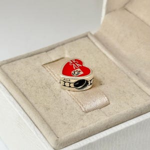 May include: A silver heart-shaped charm with red enamel and a Union Jack design. The charm has the word "ALE" inscribed on it.