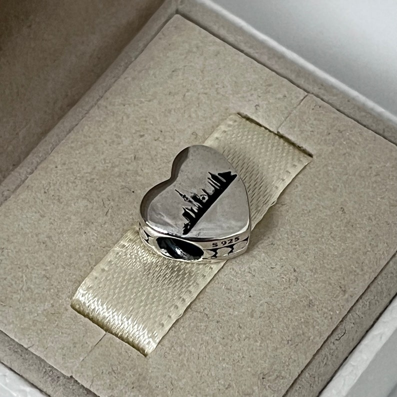 Może przedstawiać: Srebrny charm w kształcie serca z czarną sylwetką panoramy miasta. Charm jest oznaczony "S925" z boku.