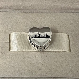 Może przedstawiać: Srebrny charm w kształcie serca z sylwetką panoramy miasta w środku. Charm ma wygrawerowane słowa "925 ALE" na boku.