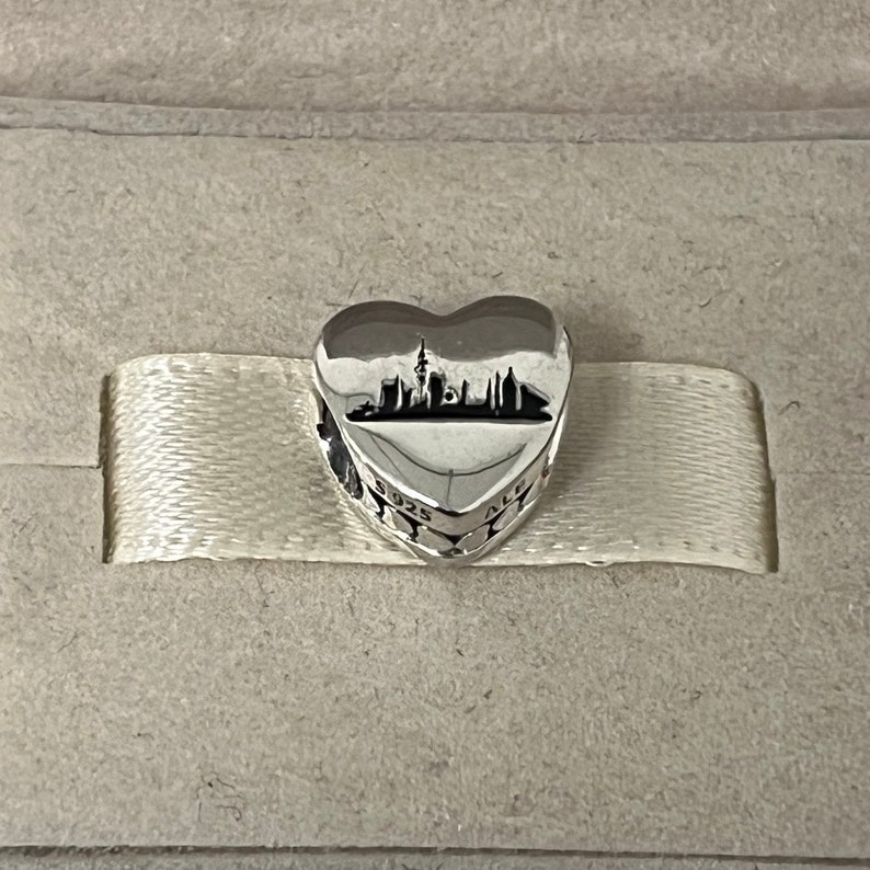 Może przedstawiać: Srebrny charm w kształcie serca z sylwetką panoramy miasta w środku. Charm ma wygrawerowane słowa "925 ALE" na boku.