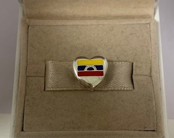 May include: A silver heart-shaped charm with a colorful enamel design of the Venezuelan flag. The charm is in a white box with a beige interior.