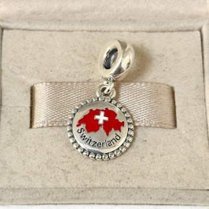 Puede incluir: Un charm de plata con un diseño de bandera suiza roja y blanca. El charm presenta el contorno de Suiza en rojo con una cruz blanca, rodeado por un borde de cuentas y la palabra "Switzerland".