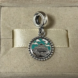 May include: Silver charm featuring a blue and white enamel design of the Folly Beach pier. The charm is round with a beaded edge and a small loop at the top.