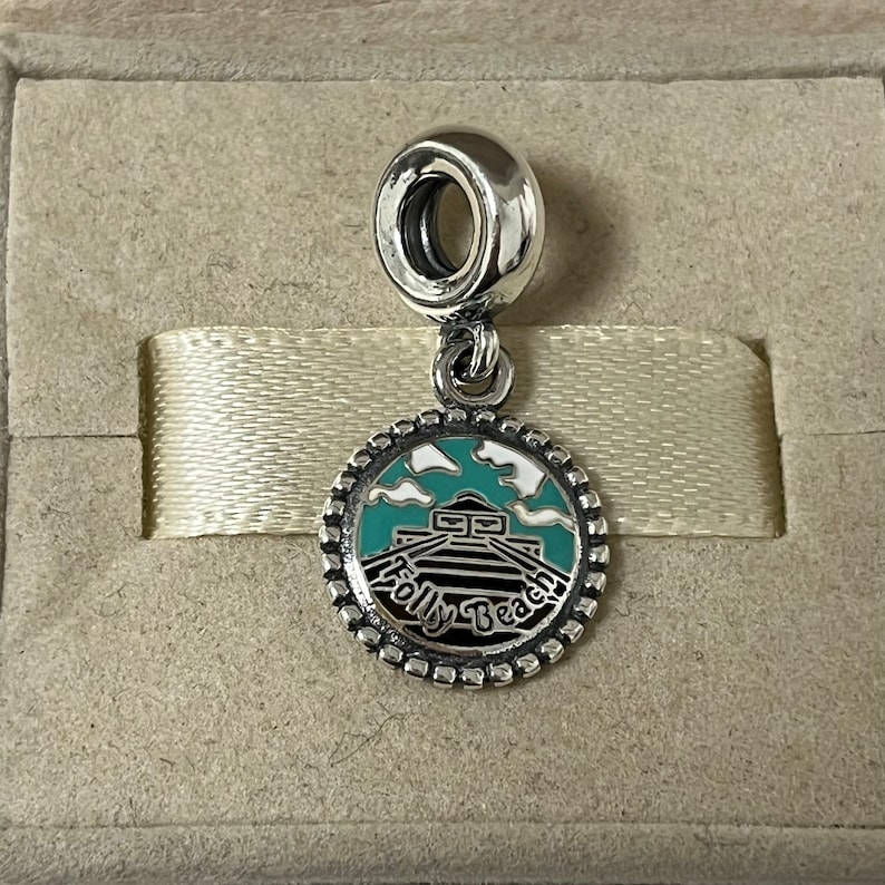 May include: Silver charm featuring a blue and white enamel design of the Folly Beach pier. The charm is round with a beaded edge and a small loop at the top.