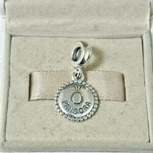 May include: A silver Pandora charm with a circular pendant. The pendant features the Pandora logo, a crown above the letter "O", and the brand name around the edge. The charm is displayed in a gray jewelry box.
