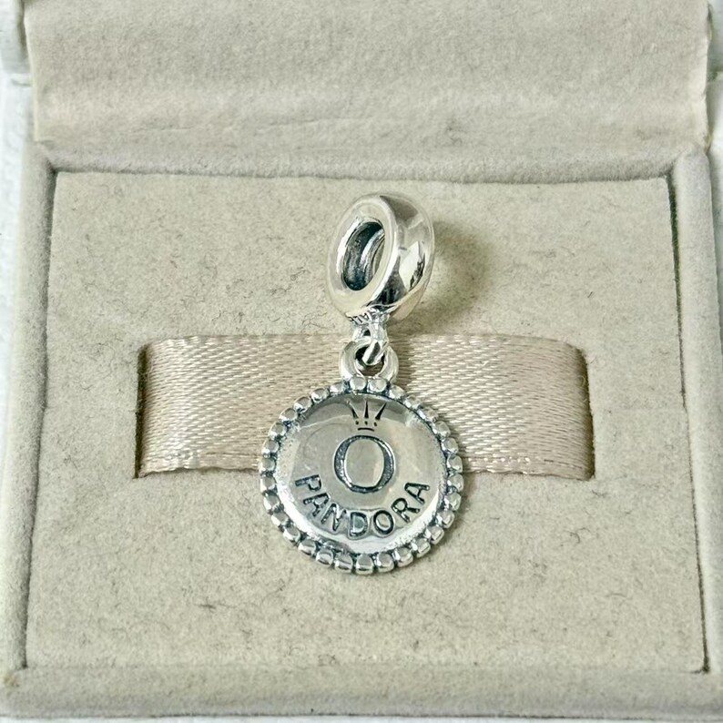 May include: A silver Pandora charm with a circular pendant. The pendant features the Pandora logo, a crown above the letter "O", and the brand name around the edge. The charm is displayed in a gray jewelry box.