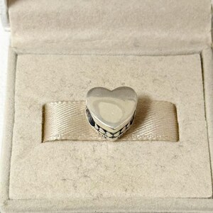 Puede incluir: Un charm de plata en forma de coraz&oacute;n con una superficie pulida. El charm tiene una banda decorativa estampada alrededor de la base y se presenta en una caja de joyer&iacute;a beige.