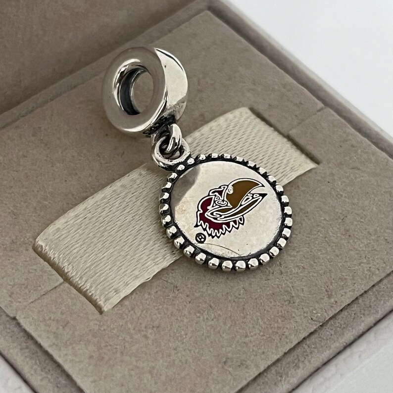 Op de afbeelding: Zilveren bedel met een rood, goud en wit logo van een vogelkop op een ronde schijf. De bedel heeft een zilveren lus aan de bovenkant.