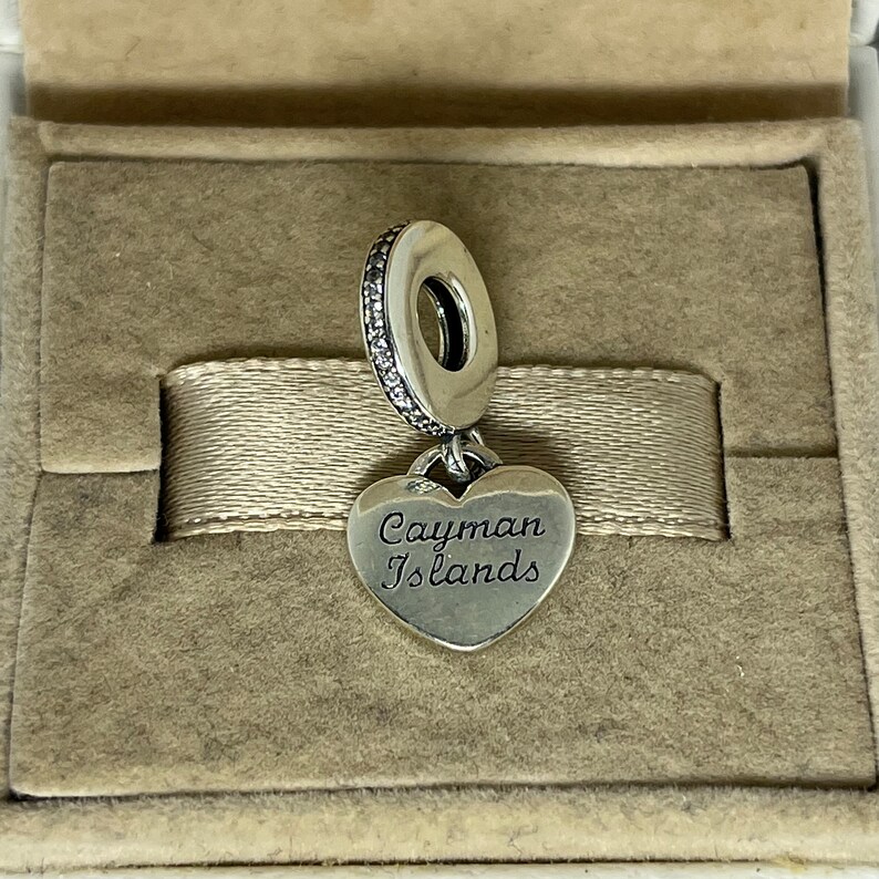 May include: Silver charm with a heart-shaped pendant that reads "Cayman Islands". The charm is in a velvet-lined box.
