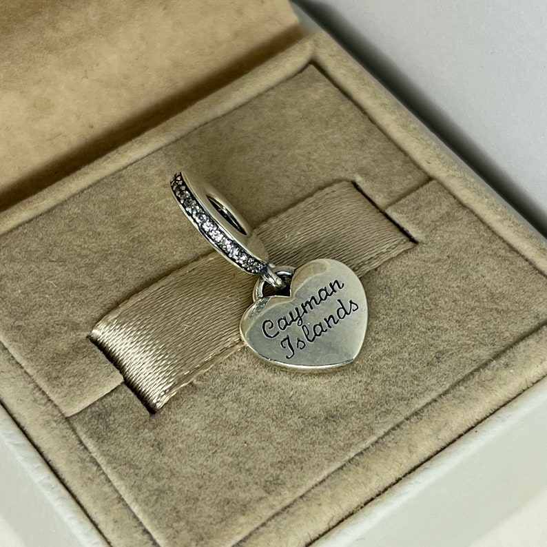 May include: Silver charm in the shape of a heart with the words "Cayman Islands" engraved on it. The charm has a silver band with clear cubic zirconia stones around the top.