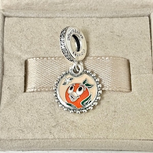 May include: Silver charm with a cartoon orange character with a green leaf and a white star on a peach background. The charm has the text "Disney" and "Disney Parks" on the back.