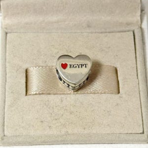 Puede incluir: Un charm plateado en forma de coraz&oacute;n con la palabra "EGYPT" en texto negro y un coraz&oacute;n rojo. El charm est&aacute; en una caja beige con una cinta beige.