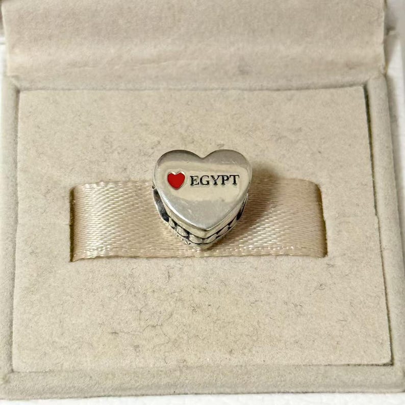 Puede incluir: Un charm plateado en forma de coraz&oacute;n con la palabra "EGYPT" en texto negro y un coraz&oacute;n rojo. El charm est&aacute; en una caja beige con una cinta beige.