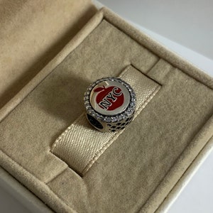 May include: A silver charm featuring a red apple design with the letters NYC in white. The charm is encircled by a ring of clear cubic zirconia stones.
