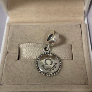 May include: A silver charm with a circular pendant featuring the word "PANDORA" and the letter "O" in the center. The charm is in a white box with a beige interior.