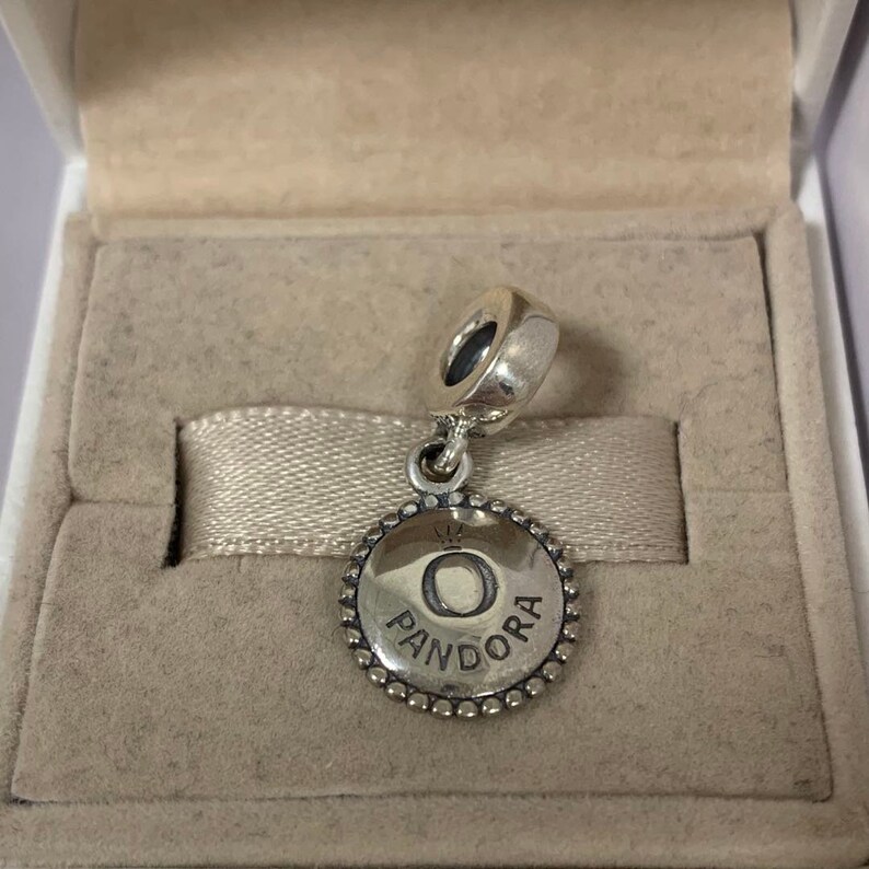 May include: A silver charm with a circular pendant featuring the word "PANDORA" and the letter "O" in the center. The charm is in a white box with a beige interior.