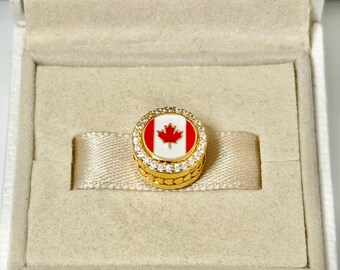 May include: A gold-colored round charm with a red and white Canadian flag design. The charm is set with small, clear gemstones around the edge and is presented in a beige box.