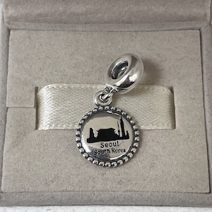 May include: Silver charm featuring a black silhouette of the Seoul skyline with the text "Seoul South Korea".