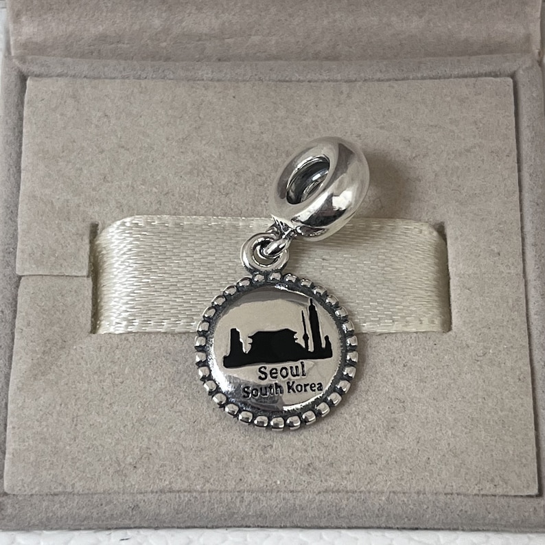 May include: Silver charm featuring a black silhouette of the Seoul skyline with the text "Seoul South Korea".