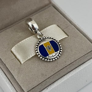 Op de afbeelding: Zilveren bedel met een blauw en geel emaille ontwerp van de vlag van Barbados. De bedel heeft een parelrand en het woord "Barbados" staat onder de vlag.