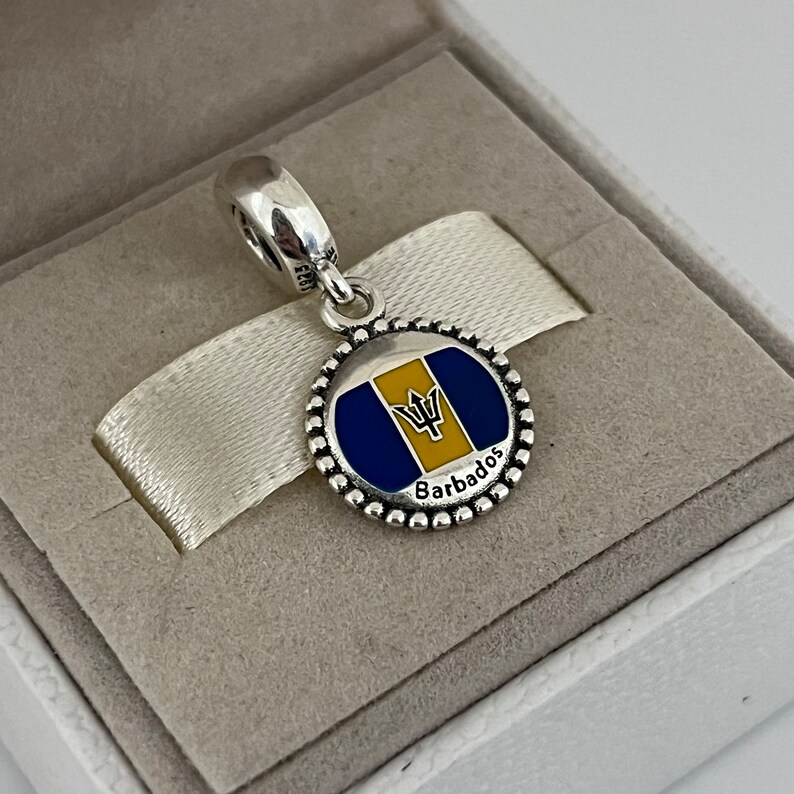 Op de afbeelding: Zilveren bedel met een blauw en geel emaille ontwerp van de vlag van Barbados. De bedel heeft een parelrand en het woord "Barbados" staat onder de vlag.