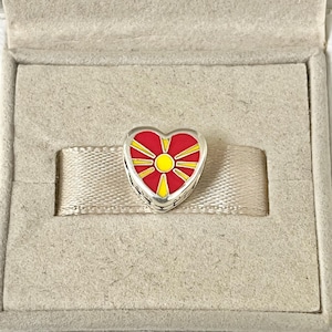 Può includere: Un ciondolo a forma di cuore in argento con un designo in smalto rosso e giallo che rappresenta la bandiera della Macedonia del Nord. Il ciondolo presenta un sole giallo con otto raggi che ne emanano, circondato da un campo rosso.
