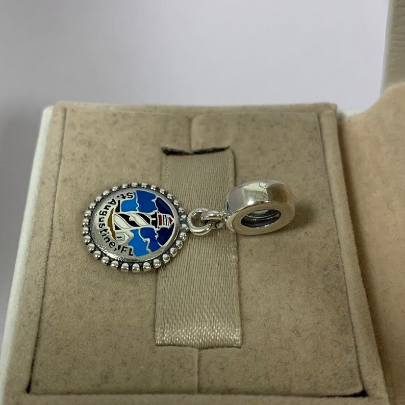 K&ouml;nnte beinhalten: Silberner Anh&auml;nger mit einem blauen und wei&szlig;en Emaille-Design eines Leuchtturms mit dem Text "St. Augustine, FL" am Rand.