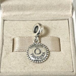 May include: A silver charm with a round pendant featuring the word "Pandora" and a crown above the letter "O".
