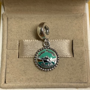 May include: Silver charm with a green and blue enamel design of a cruise ship sailing through a snowy mountain landscape. The charm has the text "Inside Passage Alaska" around the edge.