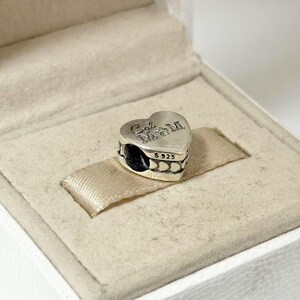 Puede incluir: Un charm plateado en forma de coraz&oacute;n con la inscripci&oacute;n "Cal M&M" y "S925". El charm se muestra en una caja blanca con una cinta de raso beige. El charm tiene un dise&ntilde;o detallado y es probablemente una joya.