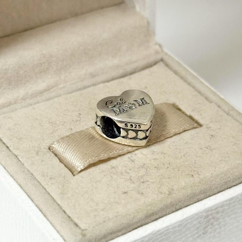 Puede incluir: Un charm plateado en forma de coraz&oacute;n con la inscripci&oacute;n "Cal M&M" y "S925". El charm se muestra en una caja blanca con una cinta de raso beige. El charm tiene un dise&ntilde;o detallado y es probablemente una joya.