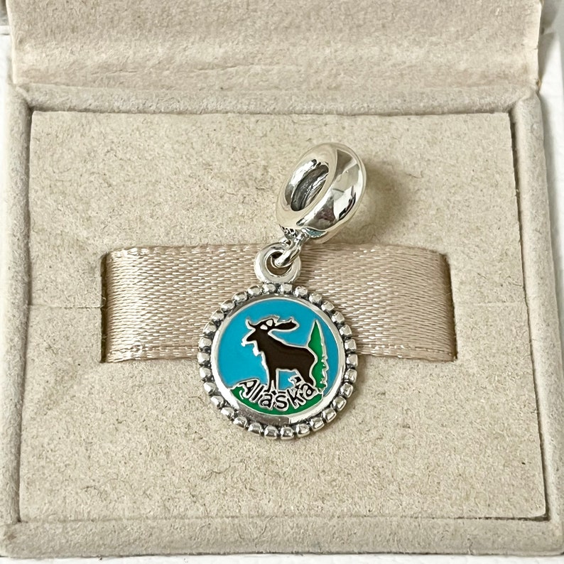 May include: Silver charm with a blue enamel circle featuring a black moose and green trees. The circle has a silver beaded border and the word "Alaska" is written in white on the bottom.