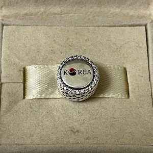 May include: A silver charm with a white and black circle design and the word "KOREA" in red letters. The charm is surrounded by a circle of clear crystals.