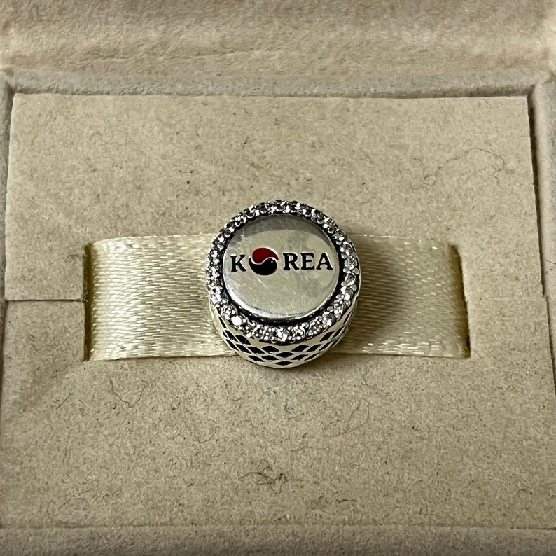 May include: A silver charm with a white and black circle design and the word "KOREA" in red letters. The charm is surrounded by a circle of clear crystals.