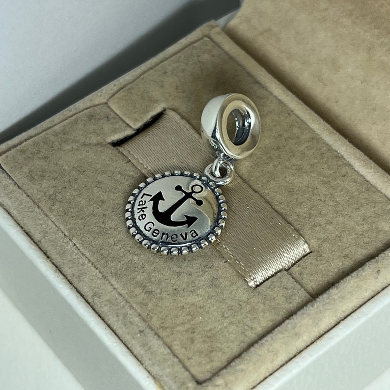 Puede incluir: Encanto de plata con un ancla negra y el texto "Lake Geneva" sobre un fondo blanco. El encanto est&aacute; en una caja blanca con un interior beige.