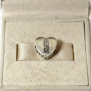 Puede incluir: Un charm de plata en forma de coraz&oacute;n con un dise&ntilde;o de Merlion, presentado en una caja beige. El charm presenta una cabeza de le&oacute;n y un cuerpo de pez, un s&iacute;mbolo de Singapur. El charm est&aacute; grabado con el texto "925 ALE".