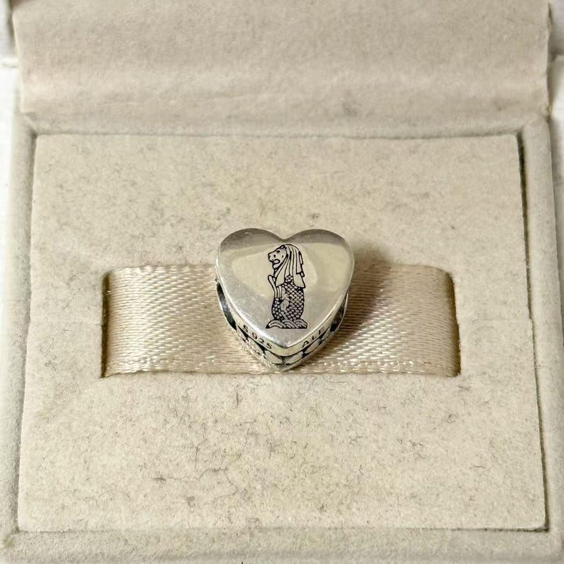 Puede incluir: Un charm de plata en forma de coraz&oacute;n con un dise&ntilde;o de Merlion, presentado en una caja beige. El charm presenta una cabeza de le&oacute;n y un cuerpo de pez, un s&iacute;mbolo de Singapur. El charm est&aacute; grabado con el texto "925 ALE".