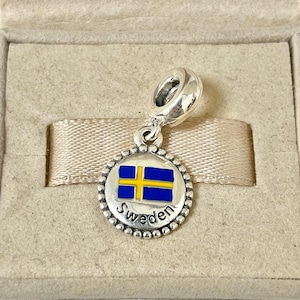 May include: A silver charm with a blue and yellow Swedish flag and the word "Sweden" inscribed around the edge.