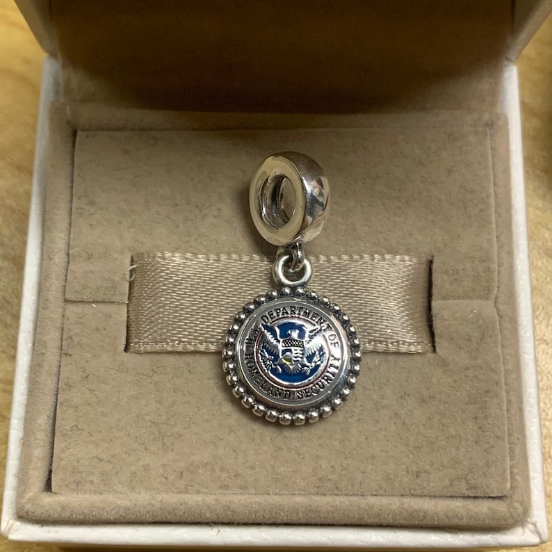 May include: Silver charm with a blue, red, and gold Department of Homeland Security seal. The charm is in a white box with a beige lining.