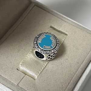 May include: A silver charm with a blue enamel circle featuring the text "Disney" and "New Orleans" around the edge. The charm is decorated with clear cubic zirconia stones.
