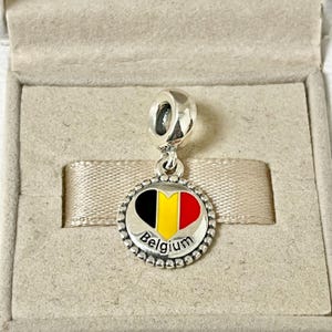Pode incluir: Berloque de prata com um design em forma de cora&ccedil;&atilde;o que apresenta as cores da bandeira belga: preto, amarelo e vermelho. O berloque tamb&eacute;m inclui a palavra "B&eacute;lgica" em letras prateadas.