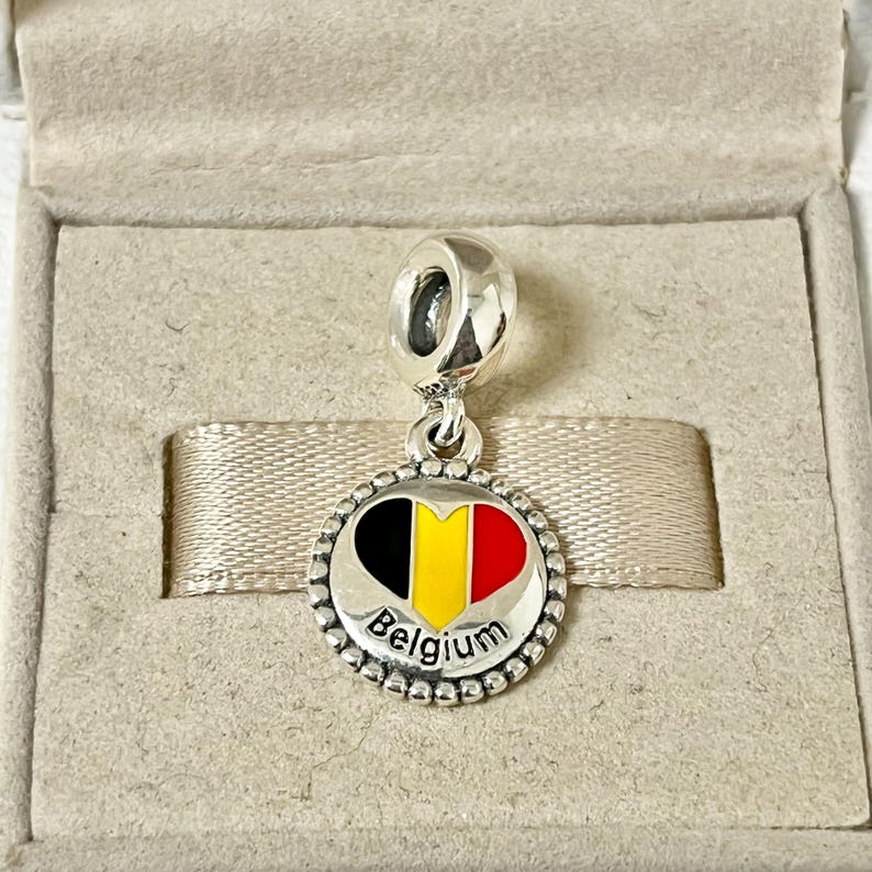 Pode incluir: Berloque de prata com um design em forma de cora&ccedil;&atilde;o que apresenta as cores da bandeira belga: preto, amarelo e vermelho. O berloque tamb&eacute;m inclui a palavra "B&eacute;lgica" em letras prateadas.