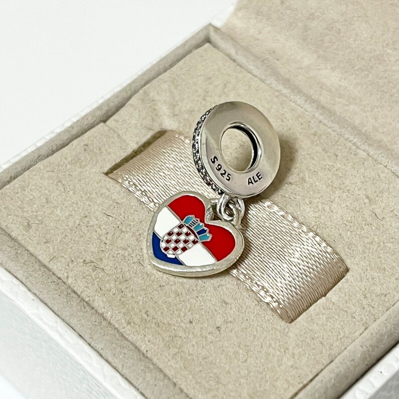 Puede incluir: Un charm de plata en forma de coraz&oacute;n con la bandera croata en &eacute;l. El charm est&aacute; unido a una cadena de plata con una cuenta redonda que dice "S925 ALE".