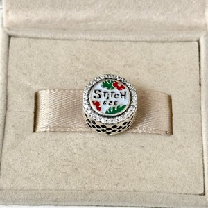 May include: A silver charm with a white background and a green and red design. The charm has the words "Stitch 626" on it and is surrounded by clear crystals.