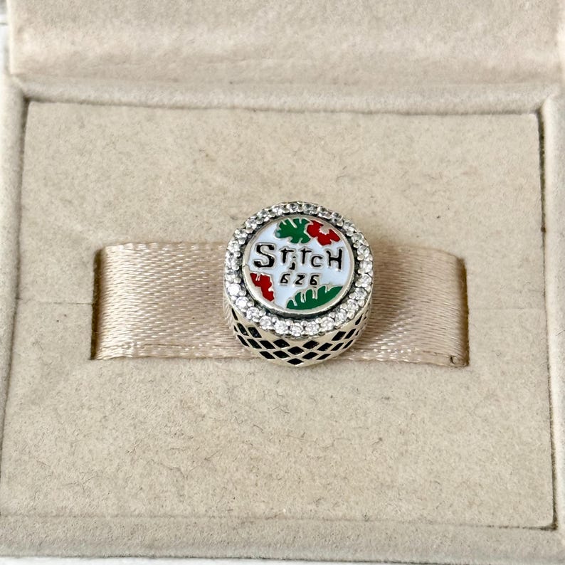 May include: A silver charm with a white background and a green and red design. The charm has the words "Stitch 626" on it and is surrounded by clear crystals.