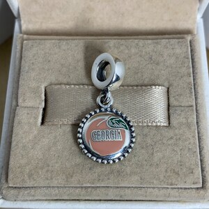 May include: Silver charm with a peach design and the word "GEORGIA" on it. The charm is in a white box with a beige interior.