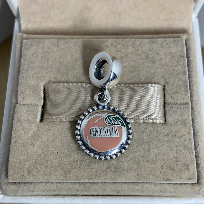 May include: Silver charm with a peach design and the word "GEORGIA" on it. The charm is in a white box with a beige interior.